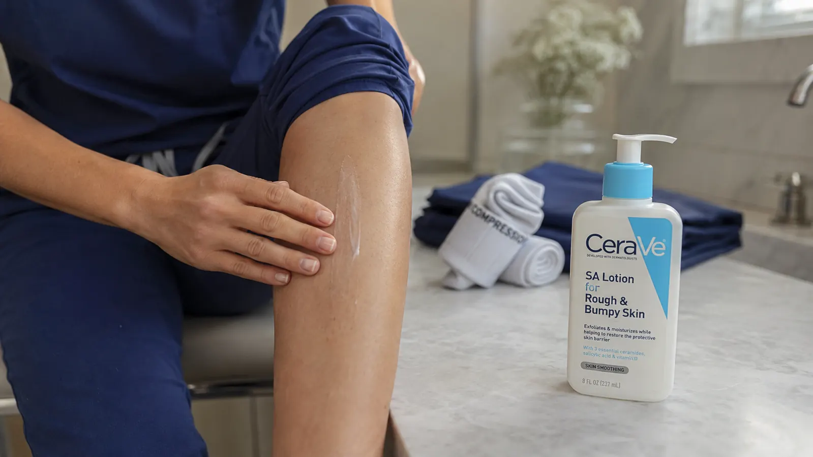 Hand smoothing white lotion onto a bare shin next to a rolled pair of compression socks on a bathroom counter