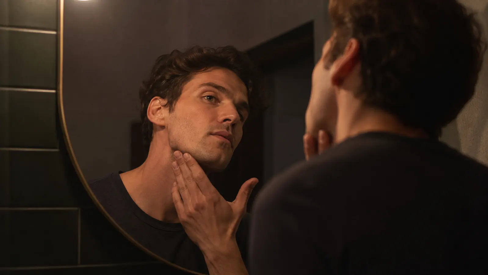Person in a bathrobe checking their chin closely in a bathroom mirror at night under warm lamp light