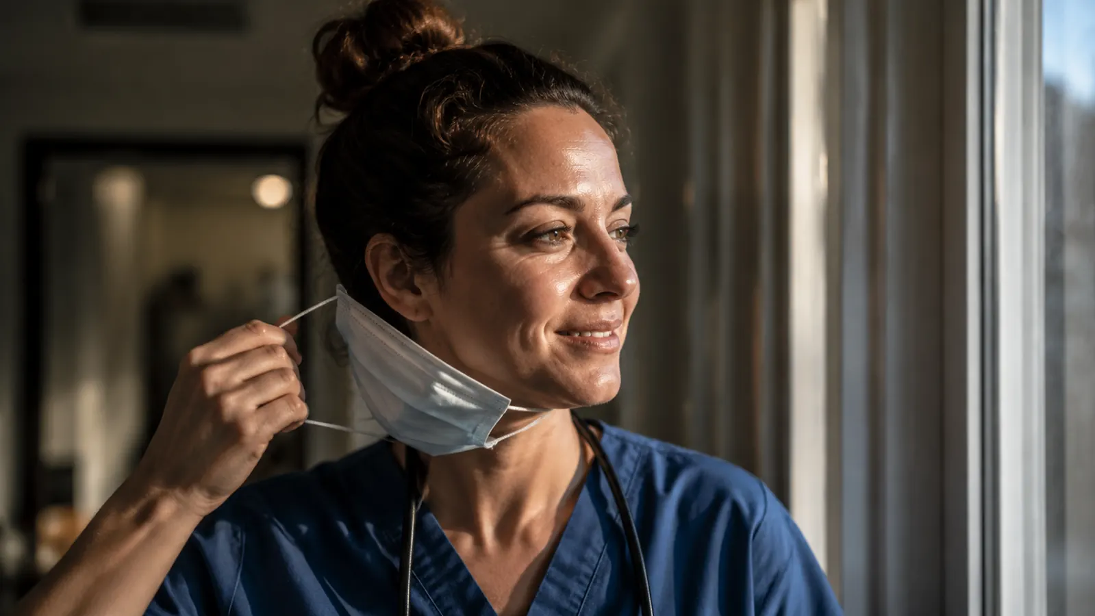 Woman removing a surgical mask at the end of a workday, standing near a window